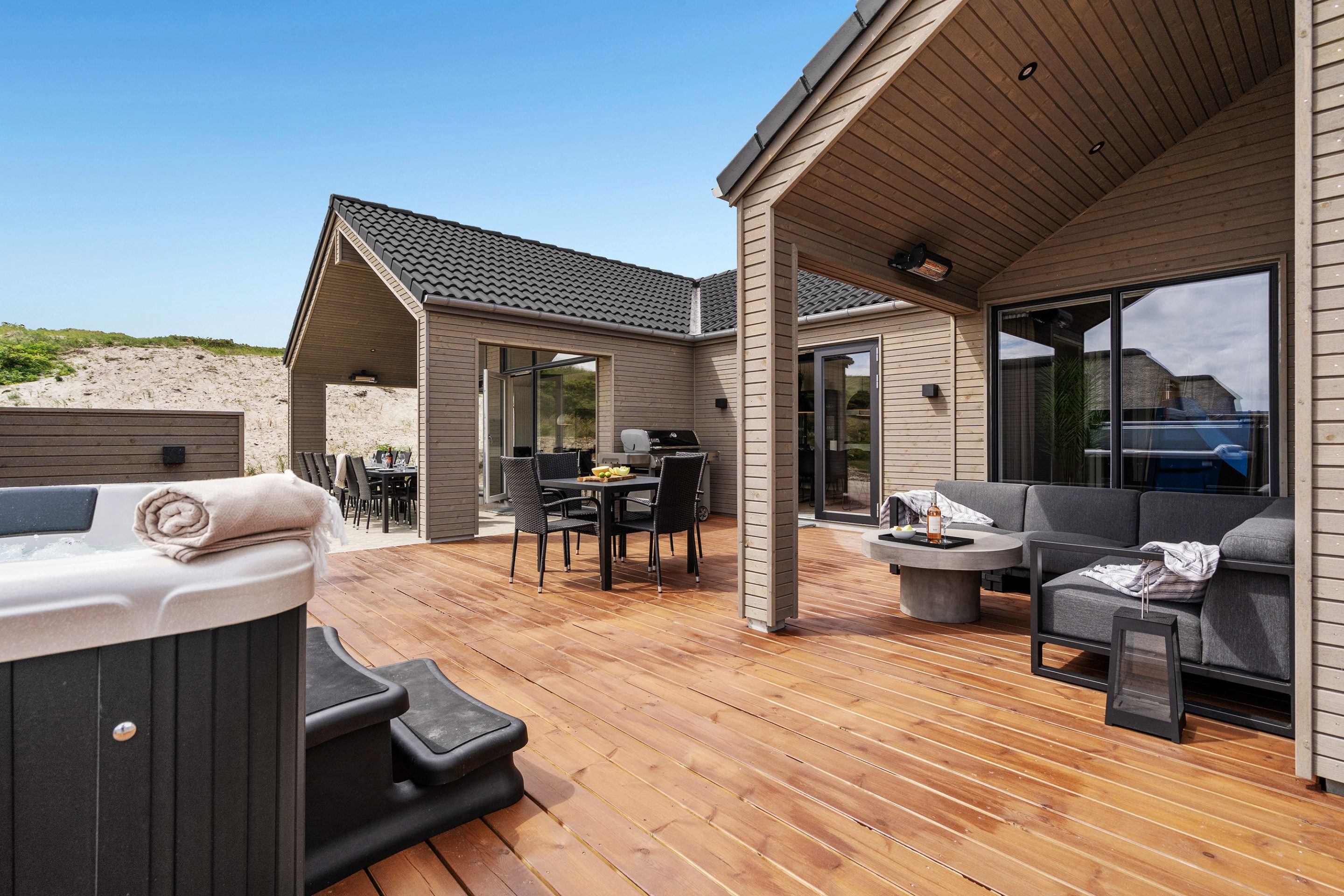 Stort skanlux sommerhus set fra terrassen. Sommerhuset er beklædt med træ og sort tegltag.